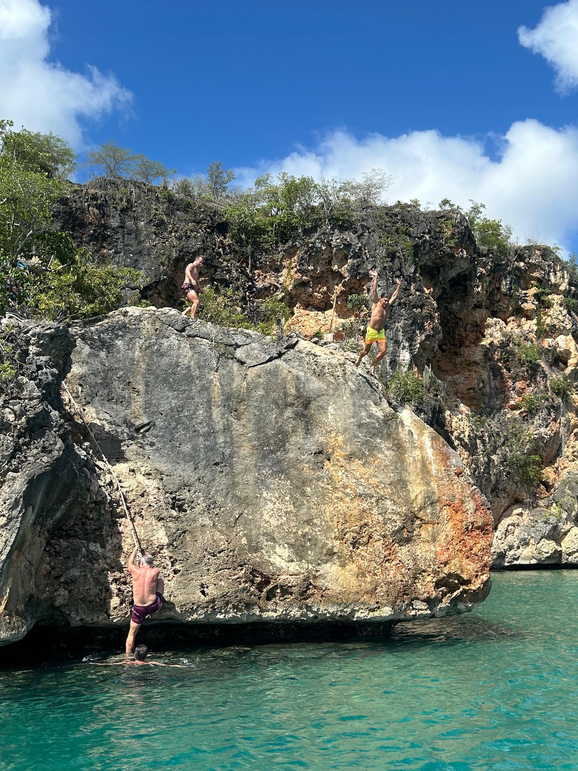 People climbing and standing on a rocky cliff above clear turquoise water on a sunny day.