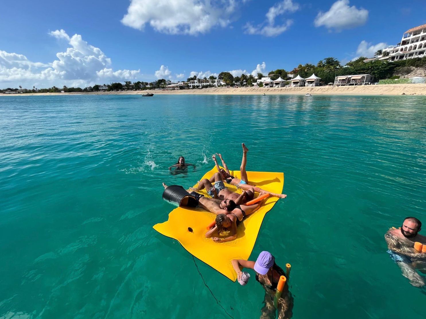 People relax on a yellow raft in turquoise water near a beach, under a blue sky with clouds.