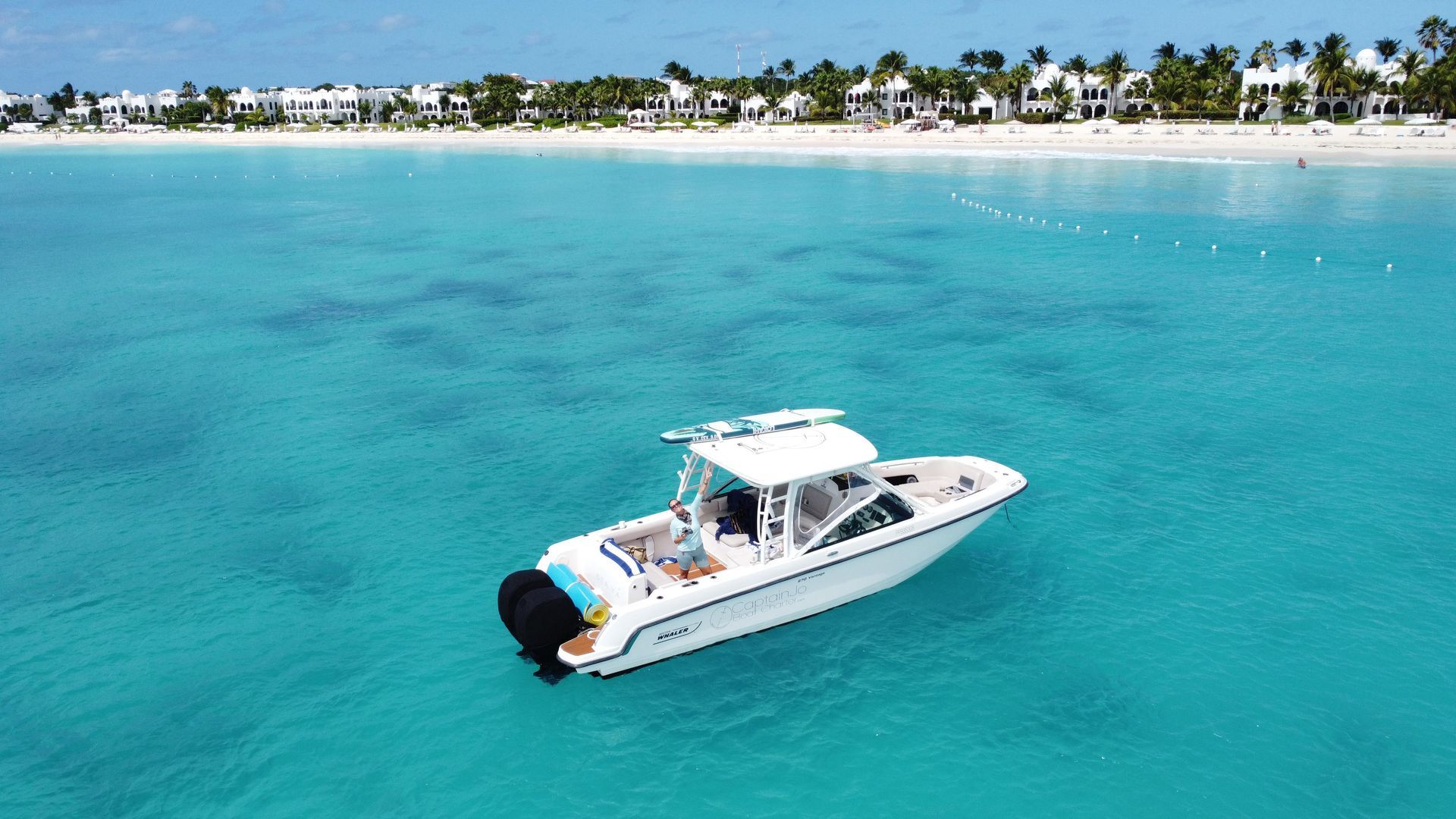 White motorboat in turquoise water near a beach with buildings and palm trees.