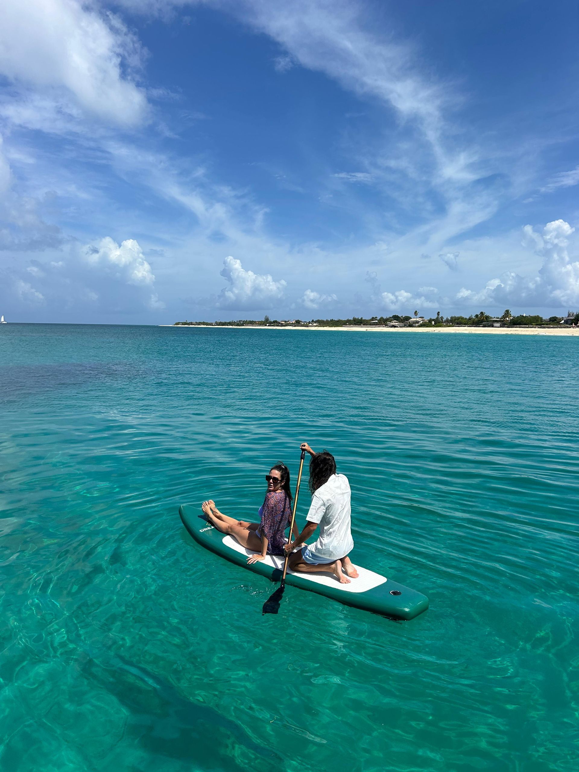 Two people on a paddleboard in turquoise water under a blue sky. One person kneels, the other sits.