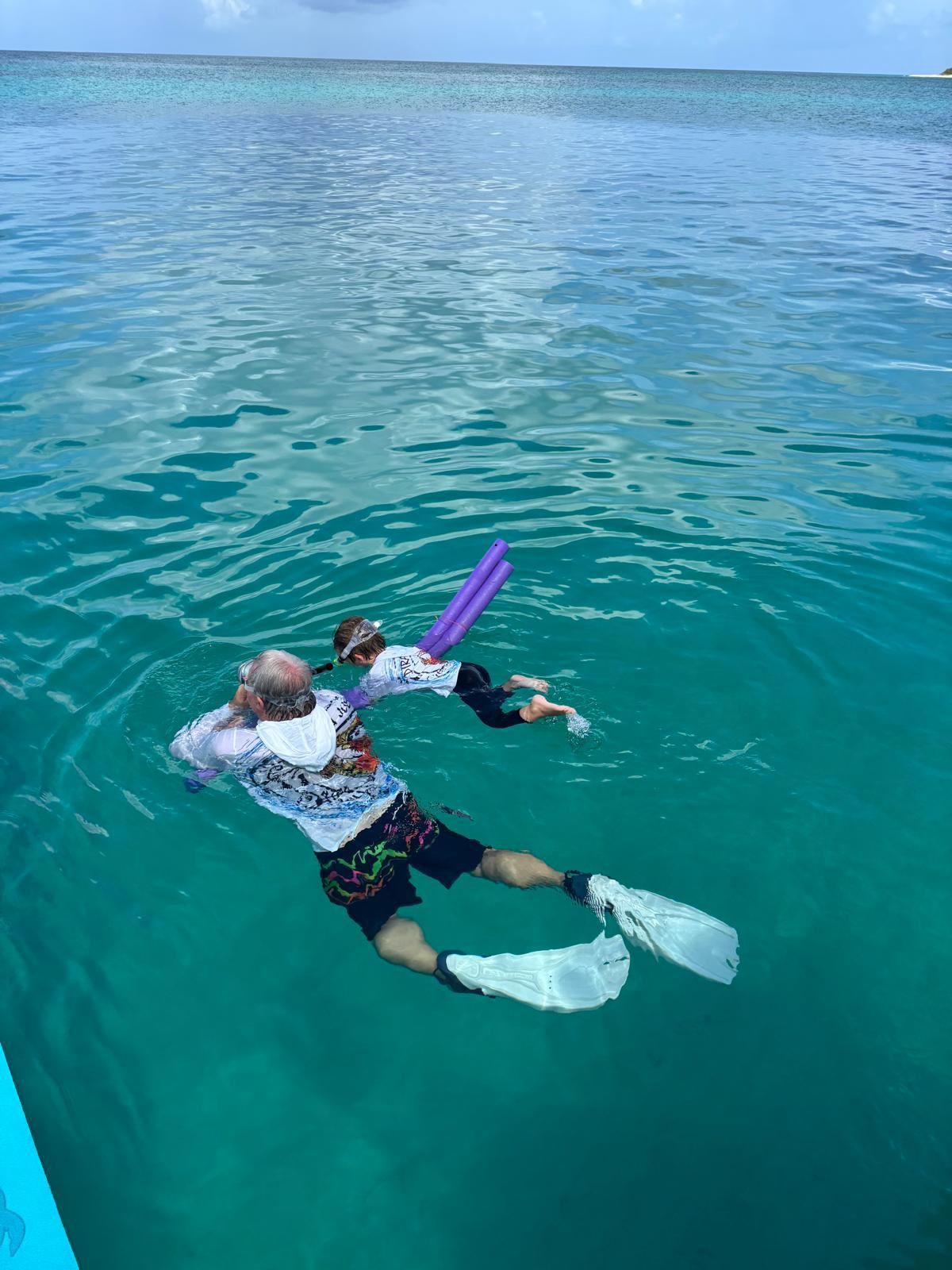 Two people snorkeling in clear turquoise water. One wears a long-sleeved shirt and flippers. The other uses a pool noodle.