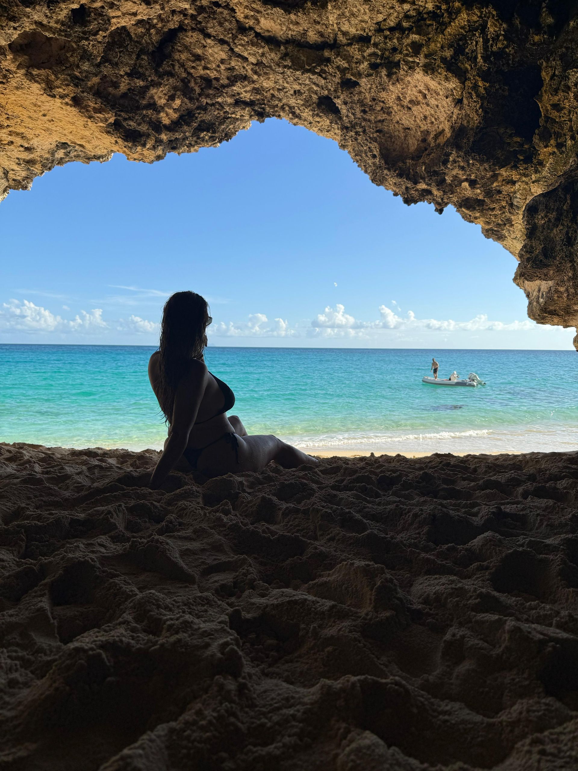 Woman seated in a cave, gazing at turquoise ocean and boat under a clear blue sky.