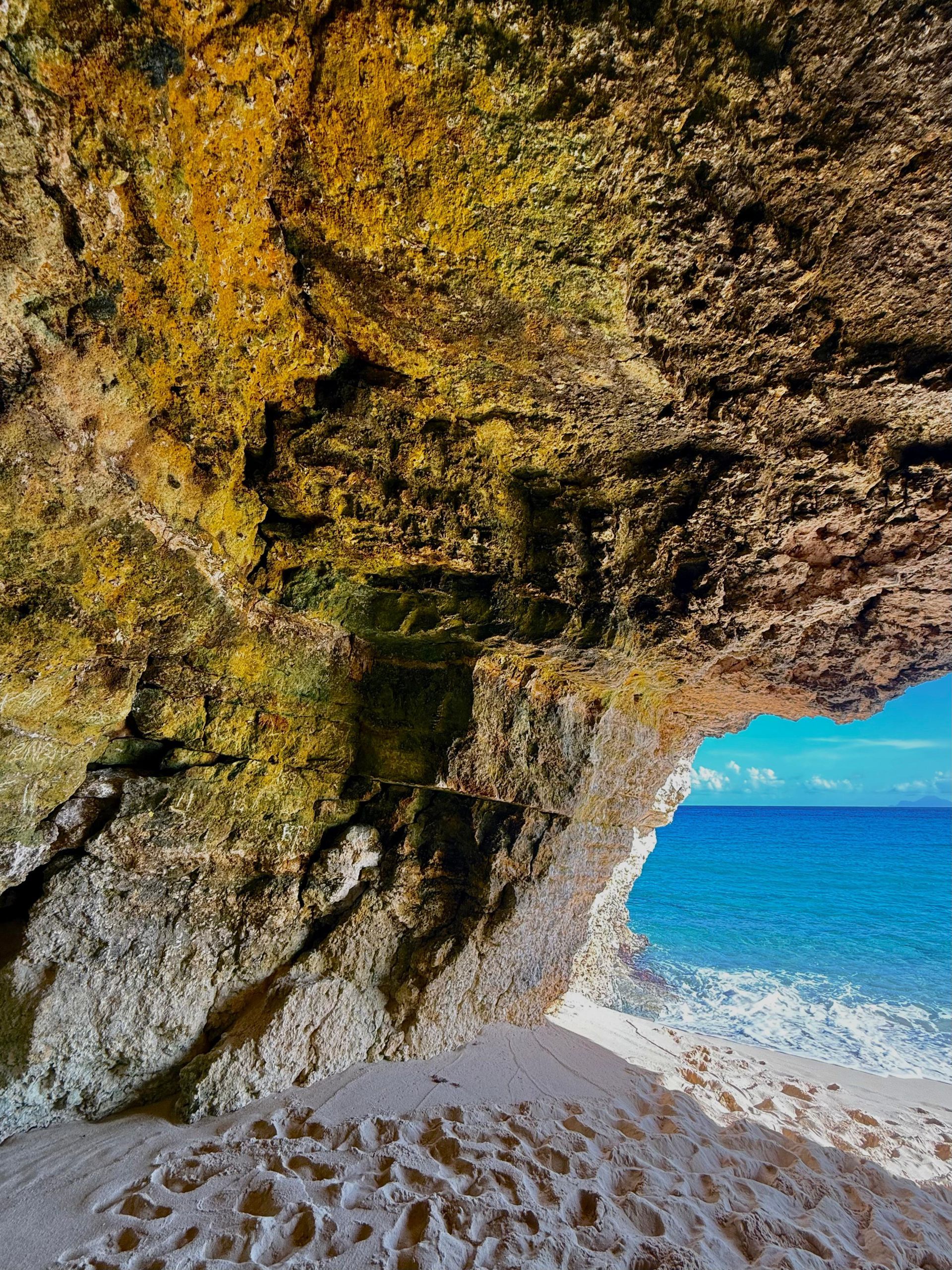 Cave opening frames a turquoise ocean view. Sandy floor, rocky ceiling with hints of yellow and green.