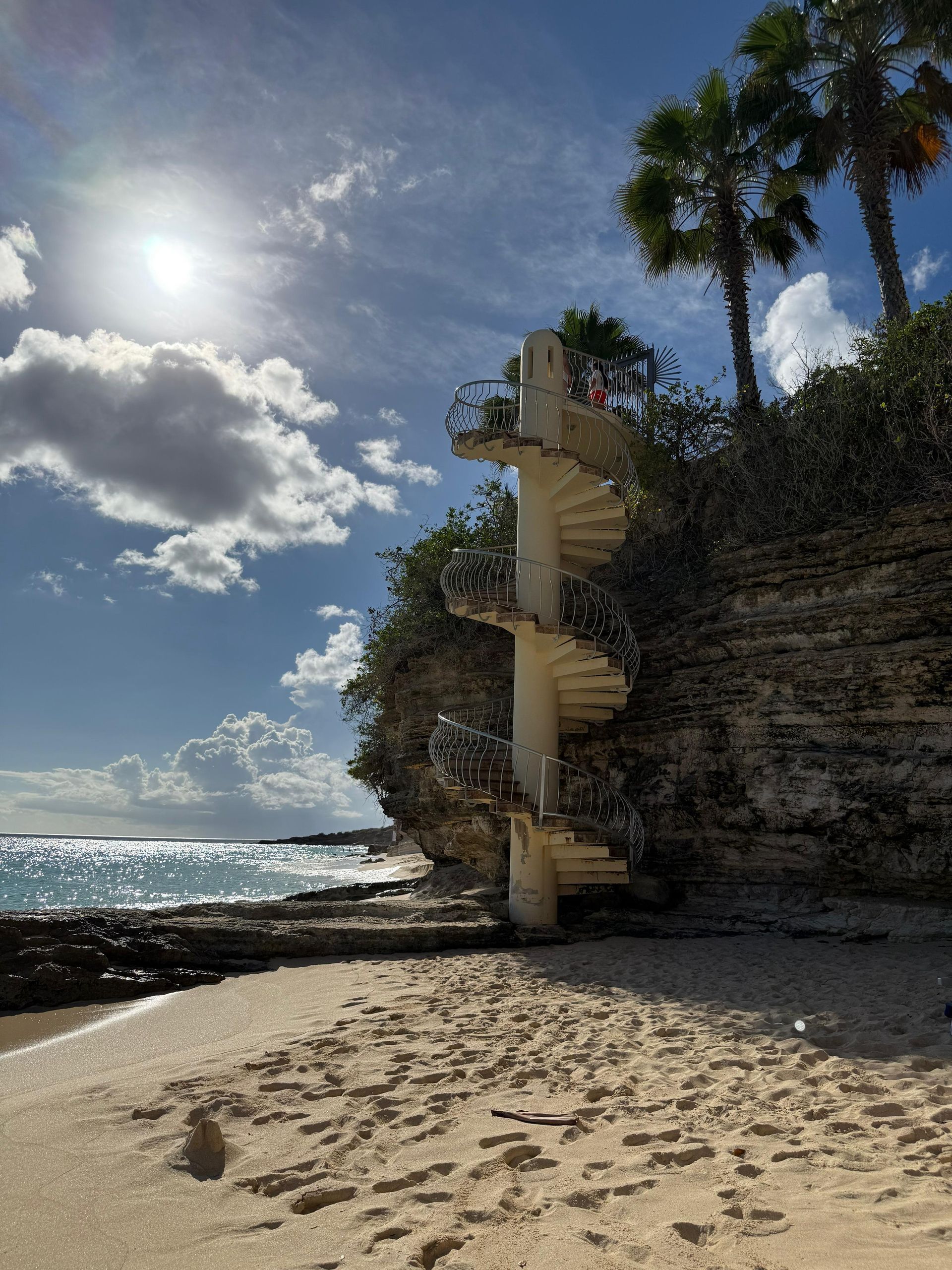 White spiral staircase on a cliff overlooking a beach and ocean under a sunny sky.