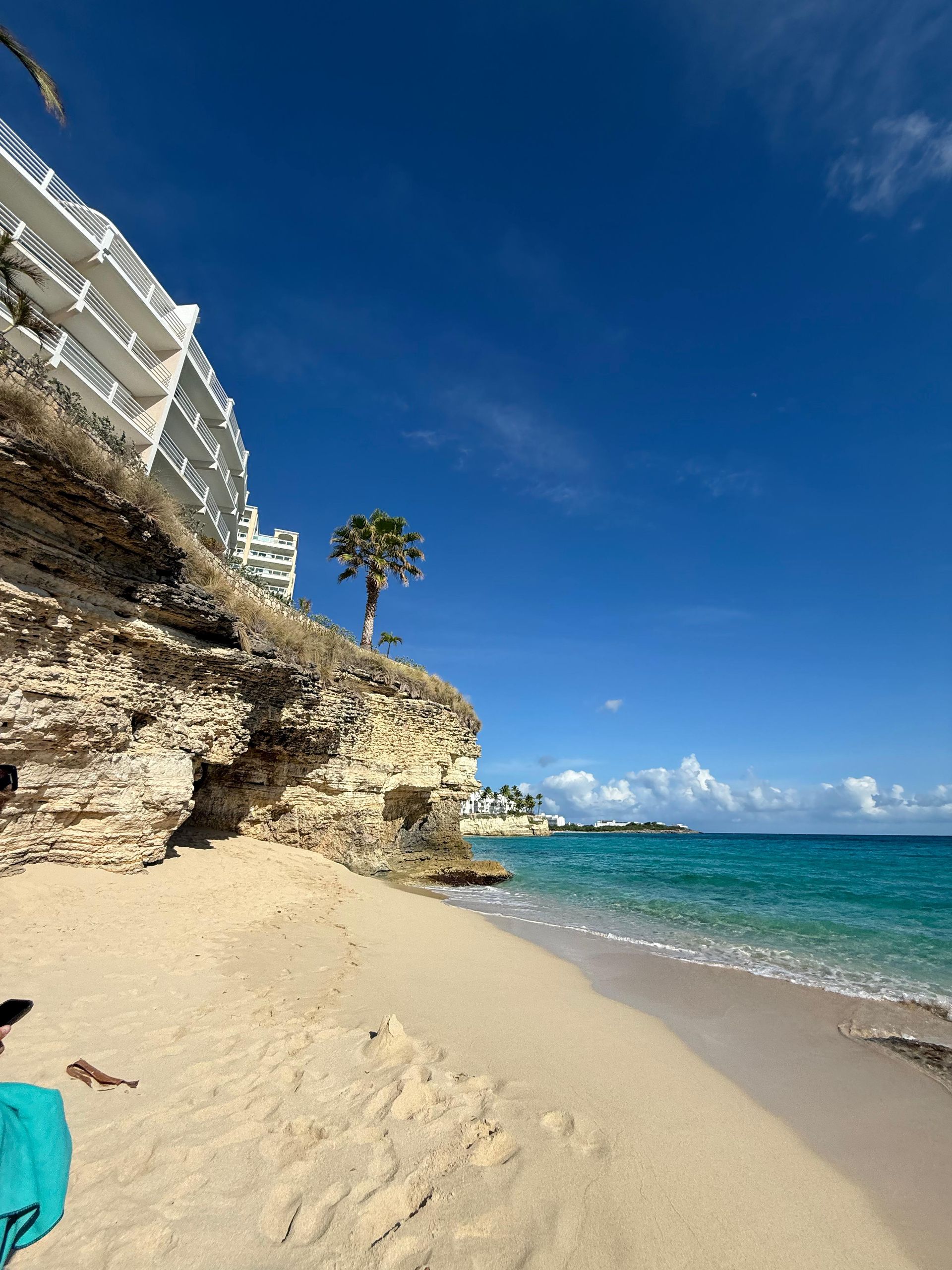 Beach scene: white sand, turquoise water, blue sky, building on cliff, palm tree.