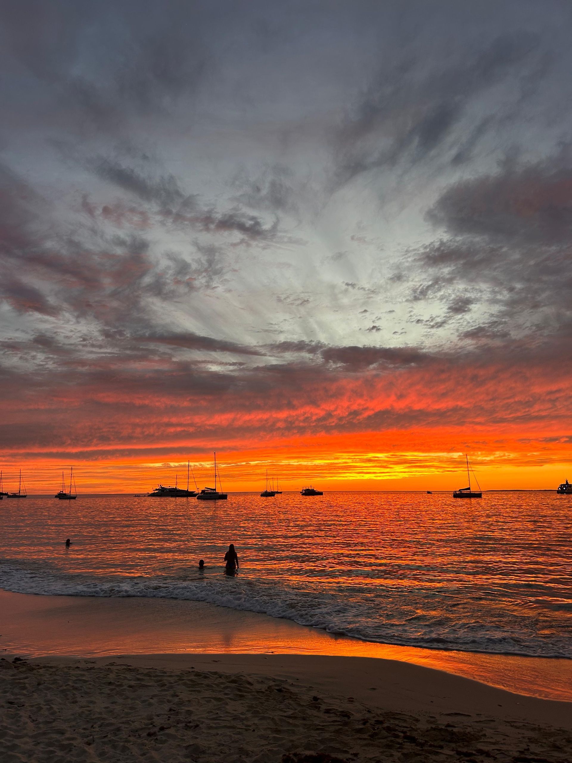 Vibrant orange and red sunset over ocean with boats silhouetted; person wading into water.