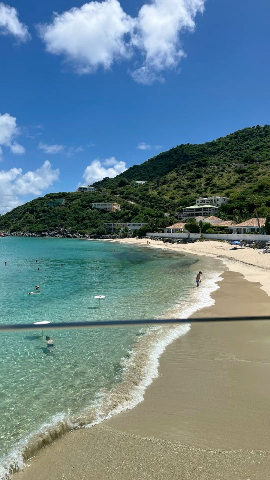 Beach with clear turquoise water, sandy shore, lush green hillside under a blue sky.