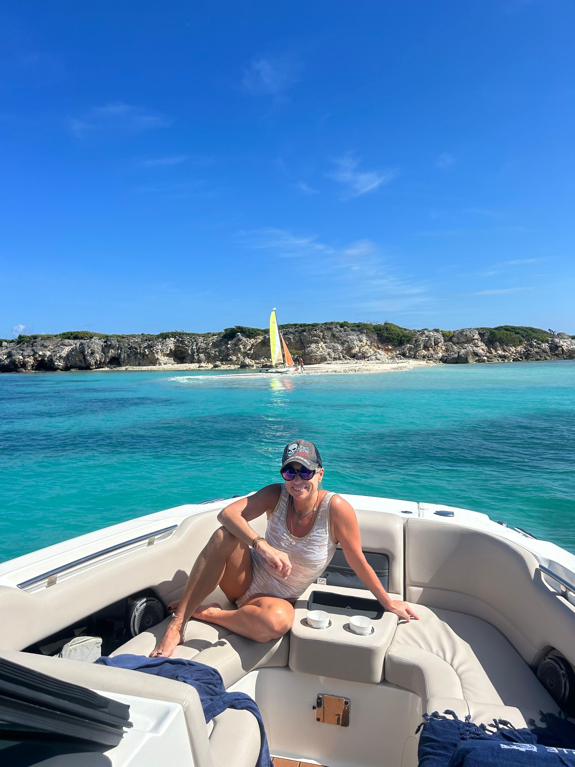 Woman on boat in turquoise water, island with lighthouse in background, clear blue sky.