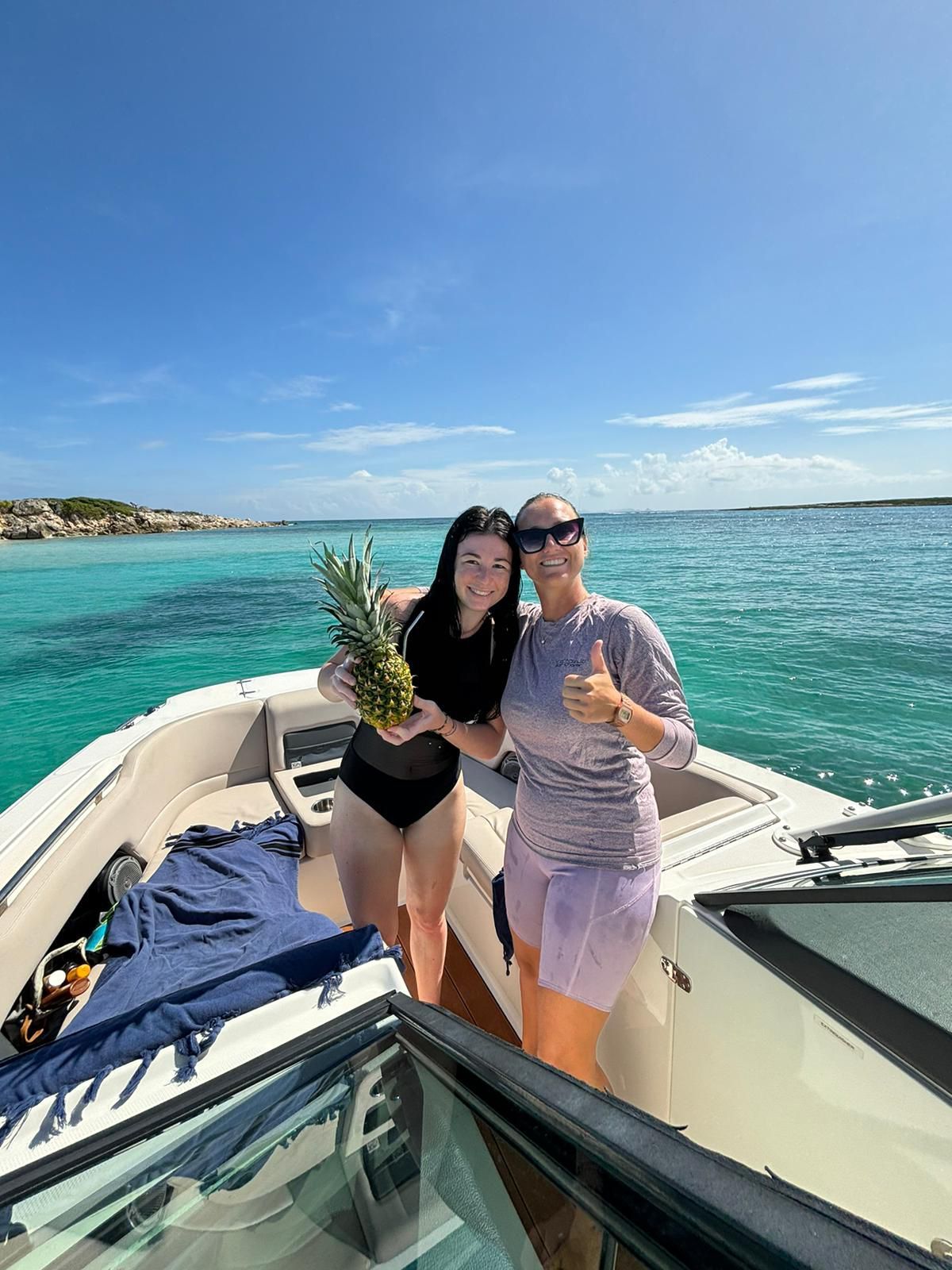 Two women on a boat; one holds a pineapple, the other gives a thumbs-up, with turquoise water and blue sky.