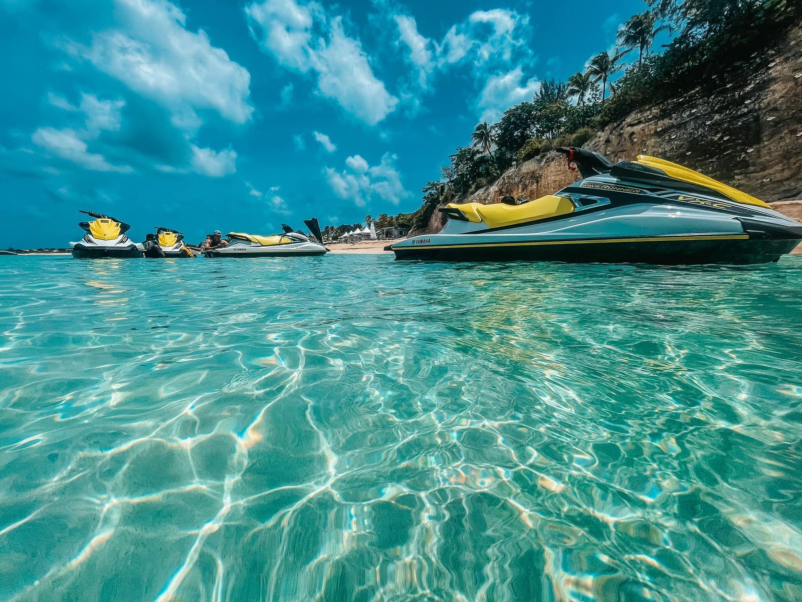 Jet skis in clear turquoise water near a beach, under a partly cloudy blue sky.