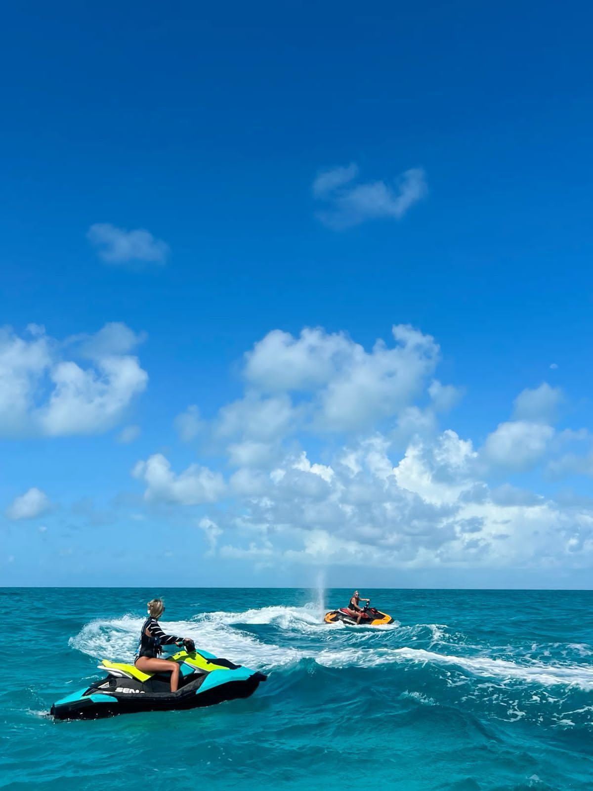 Two people on jet skis in turquoise water under a partly cloudy blue sky.