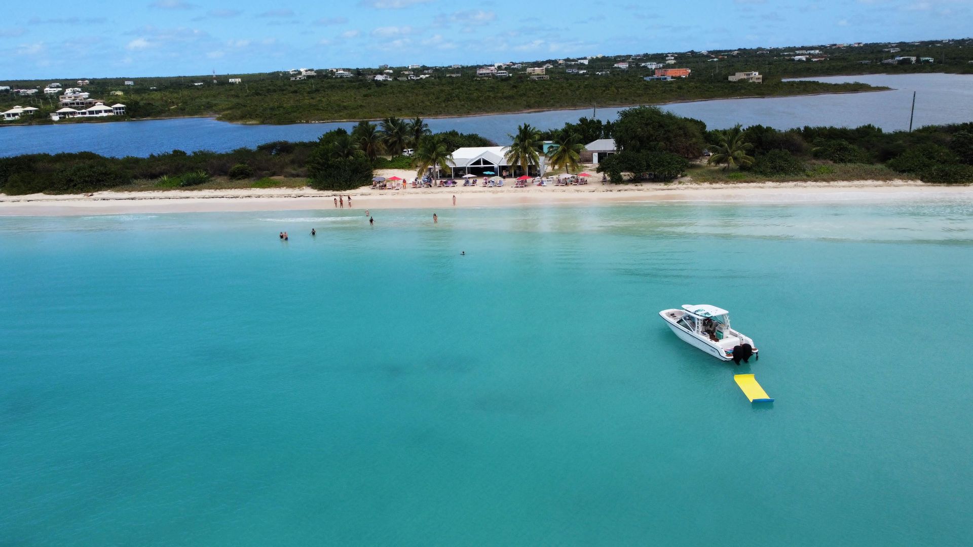 A boat floats on turquoise water near a white sandy beach. Buildings and trees line the shore.