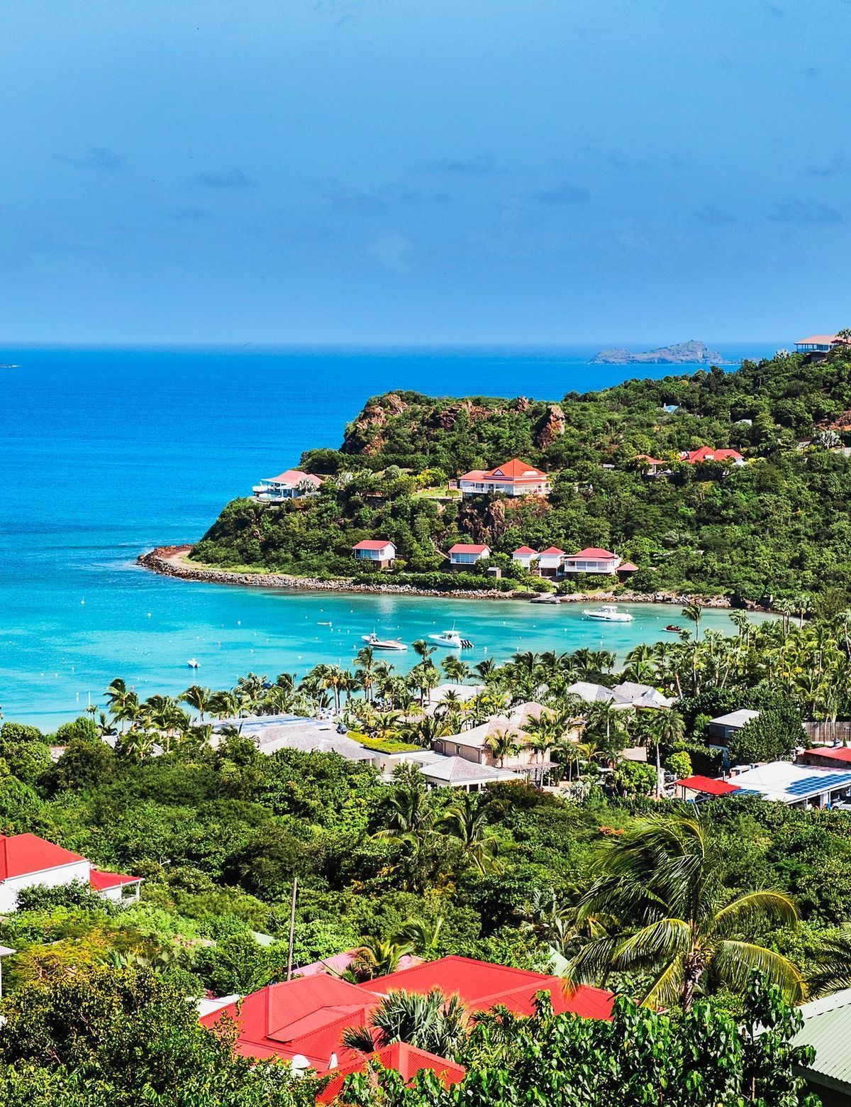 Tropical coastal view with turquoise water, green hills, red-roofed villas, and a clear blue sky.