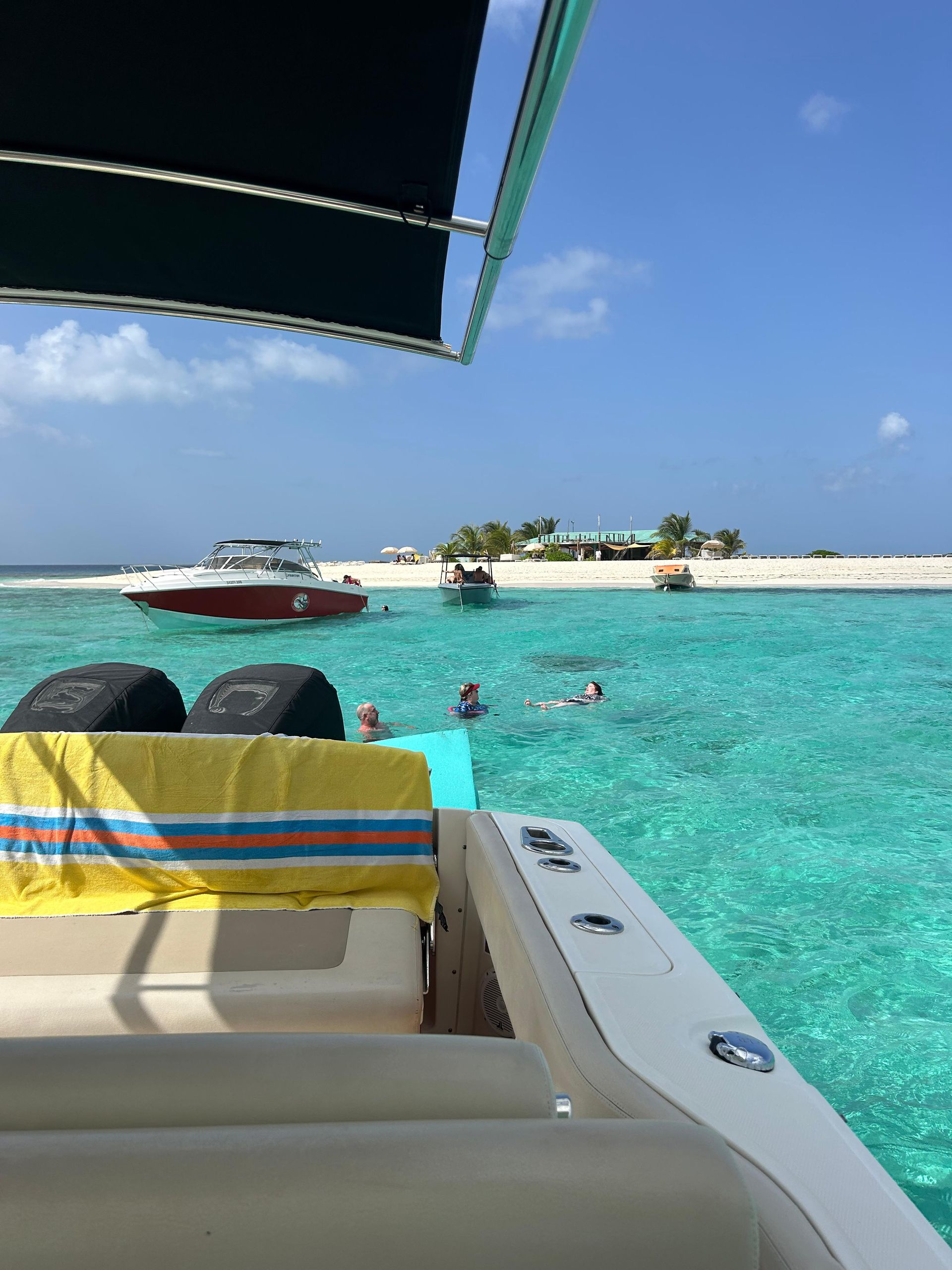 Boat overlooking turquoise water with swimmers, other boats, and a sandy island under a sunny sky.