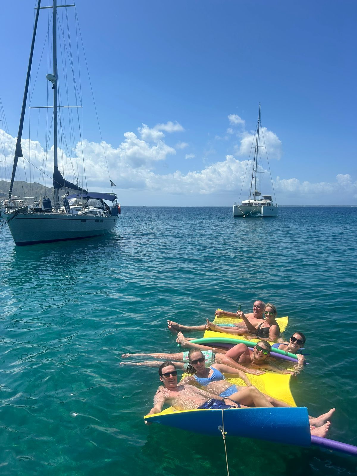 People on a yellow raft in turquoise water; sailboats in the background under a blue sky.