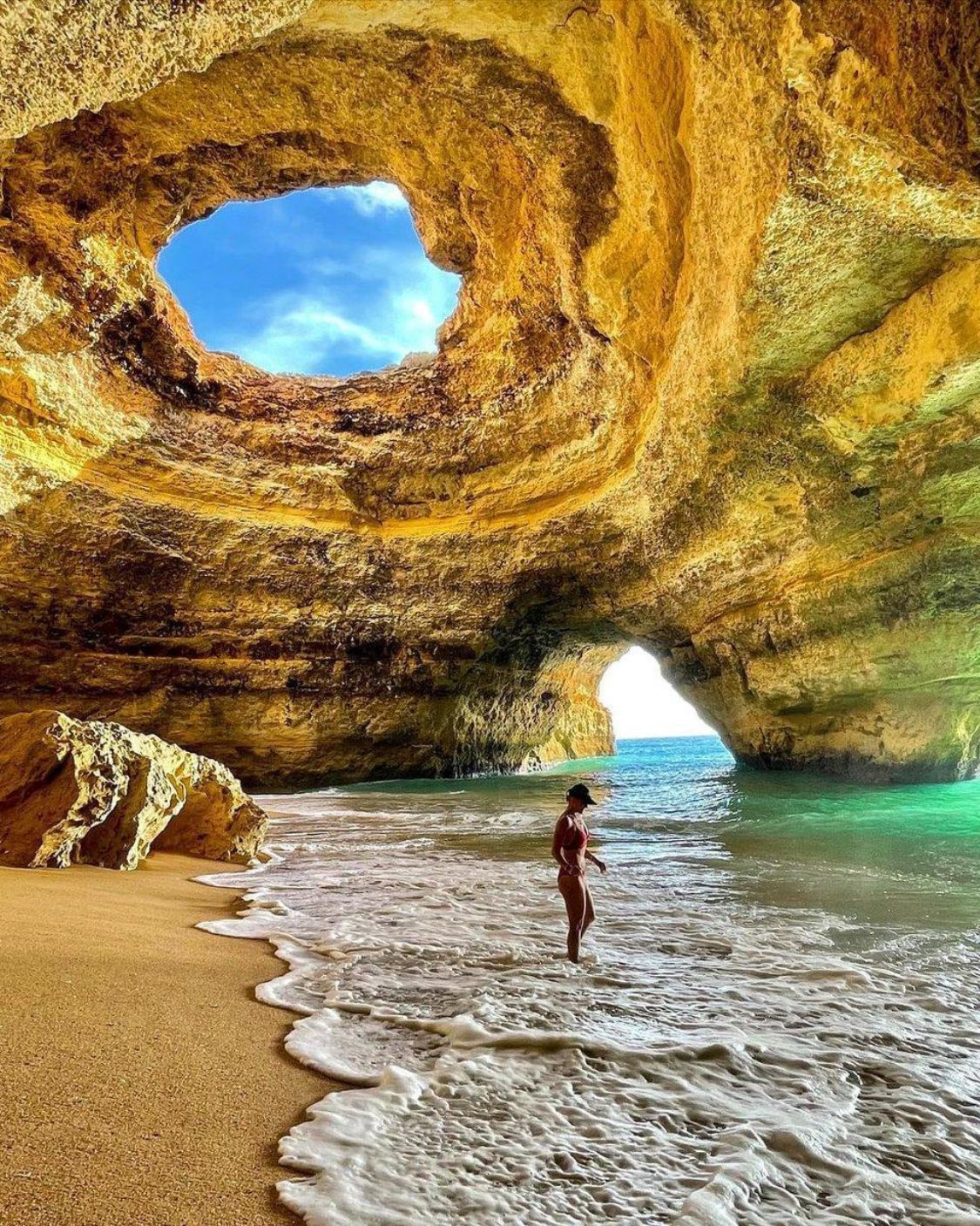 Woman wading in turquoise water inside a golden-hued sea cave with an open skylight, Portugal.