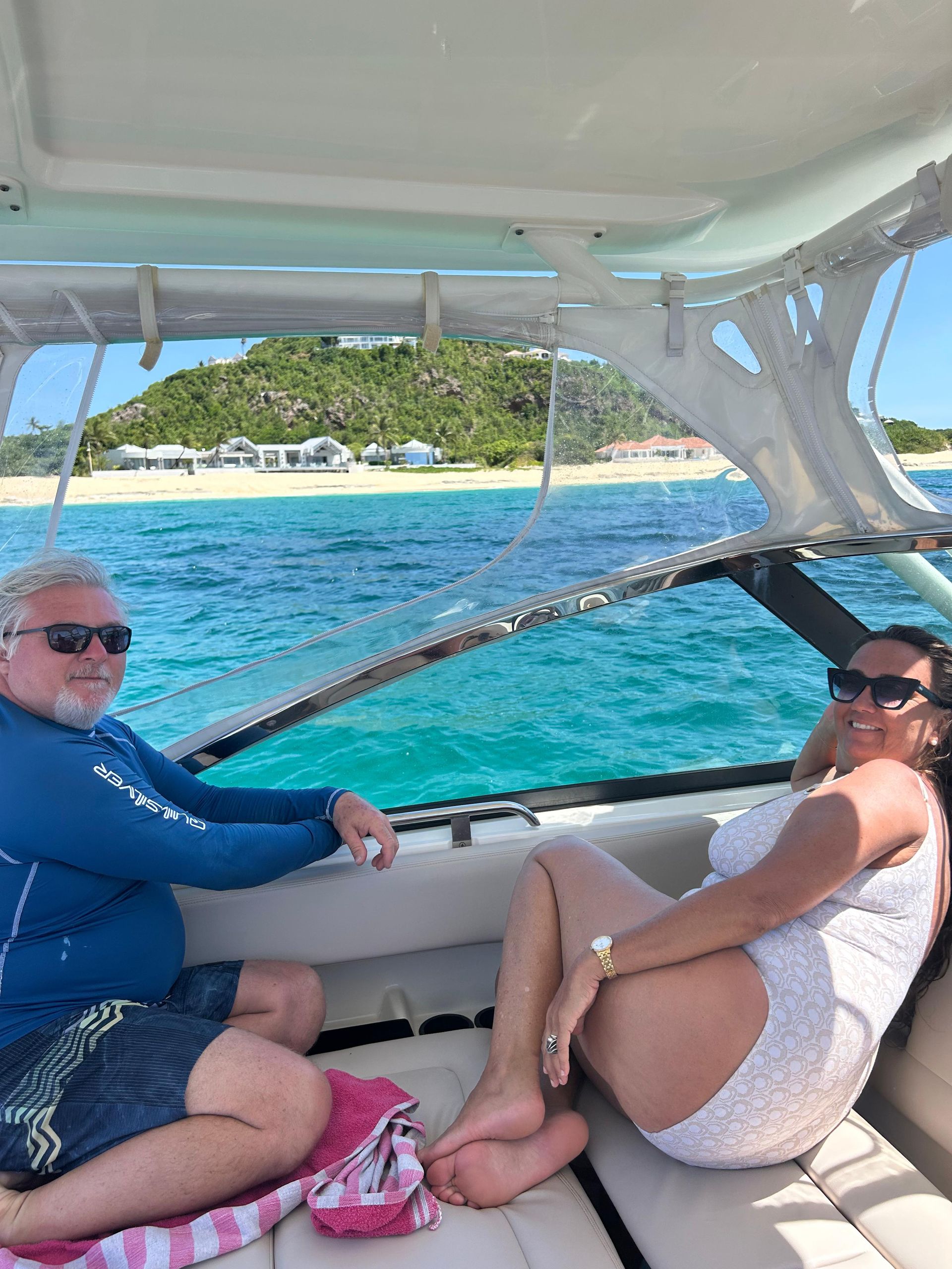 Couple on a boat in turquoise water, sunny day. Man in blue shirt, woman in white dress, beachfront in background.