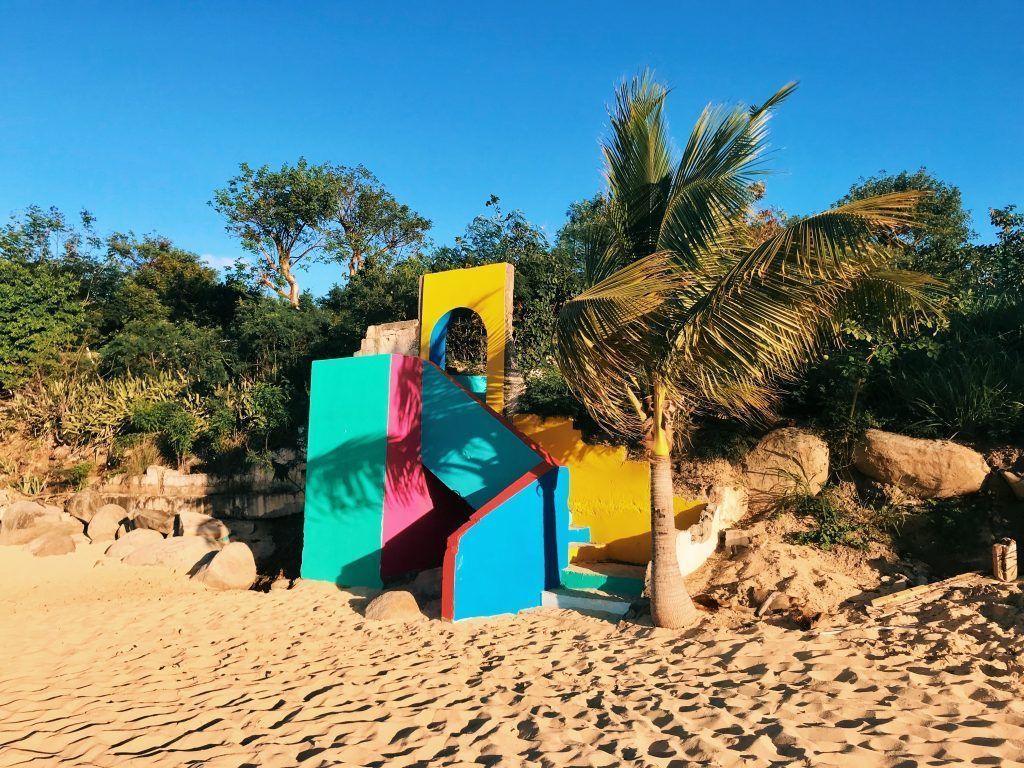 Colorful geometric sculpture on a beach with palm tree, set against a backdrop of greenery under blue sky.