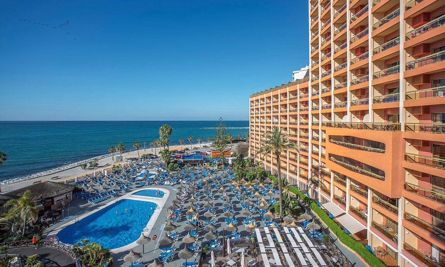 Seaside resort with blue pools, sun loungers, and a tall orange hotel beside the ocean