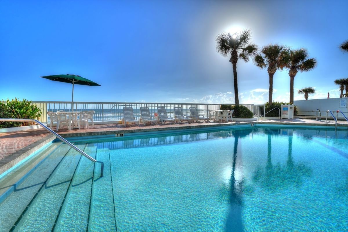 Oceanfront pool with turquoise water, white lounge chairs, palm trees, and ocean views under a bright sky