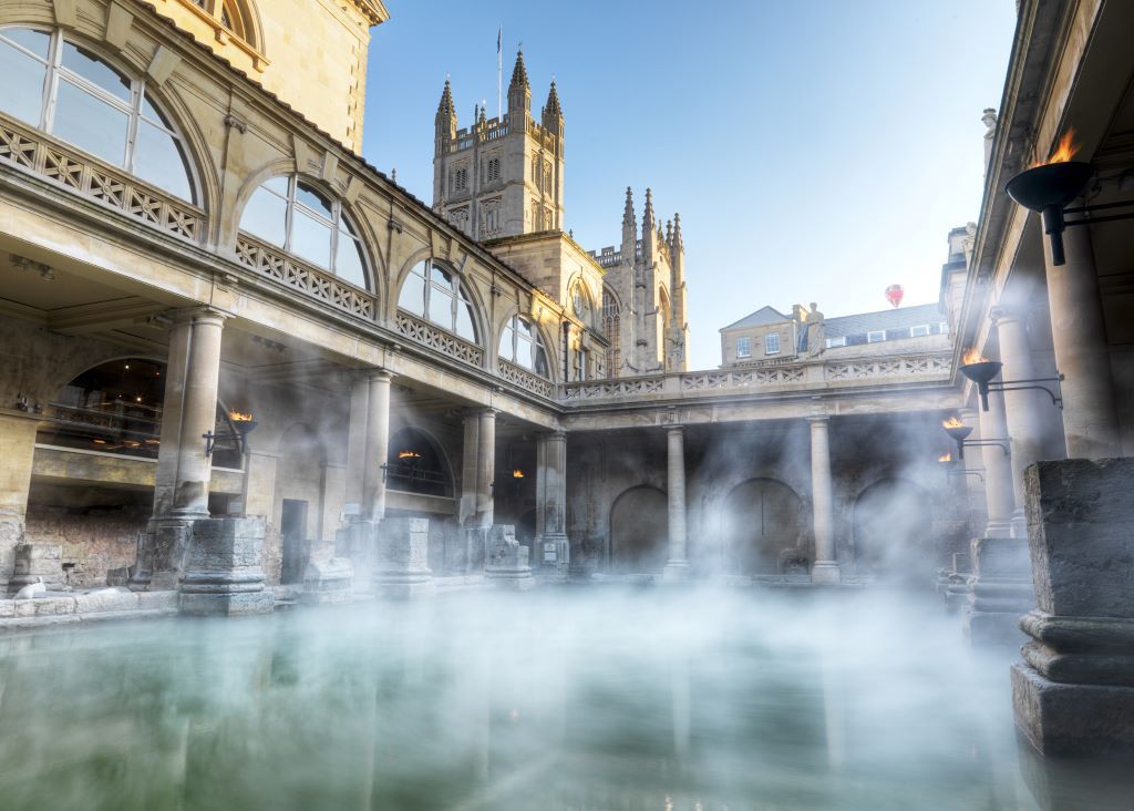 Roman Baths in Bath, England, with steaming water, arched walkways, and a towering cathedral in the background.