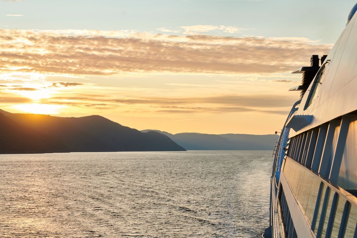 Cruise ship sailing on calm water toward mountains at sunset.