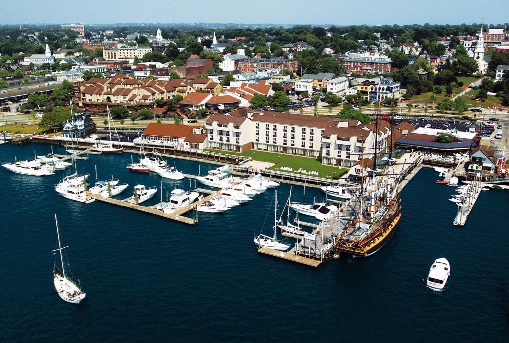 Aerial view of a harbor with boats and a tall ship, buildings and town in the background, blue water and sky.
