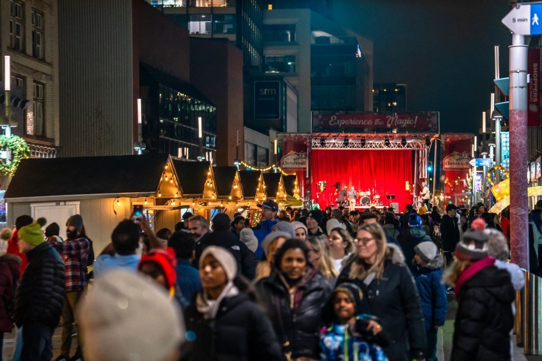 Crowded nighttime city street market with people walking toward a brightly lit red stage and festive lights