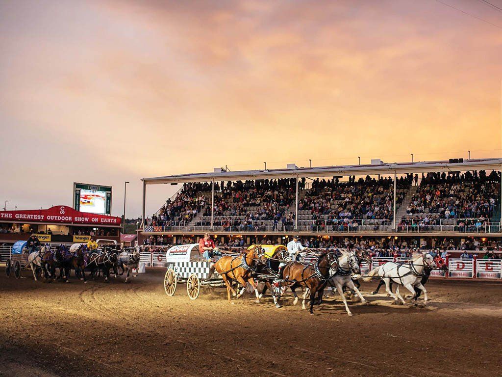 Wagon race in arena, horses pulling wagons. Crowded stands under a sunset sky.