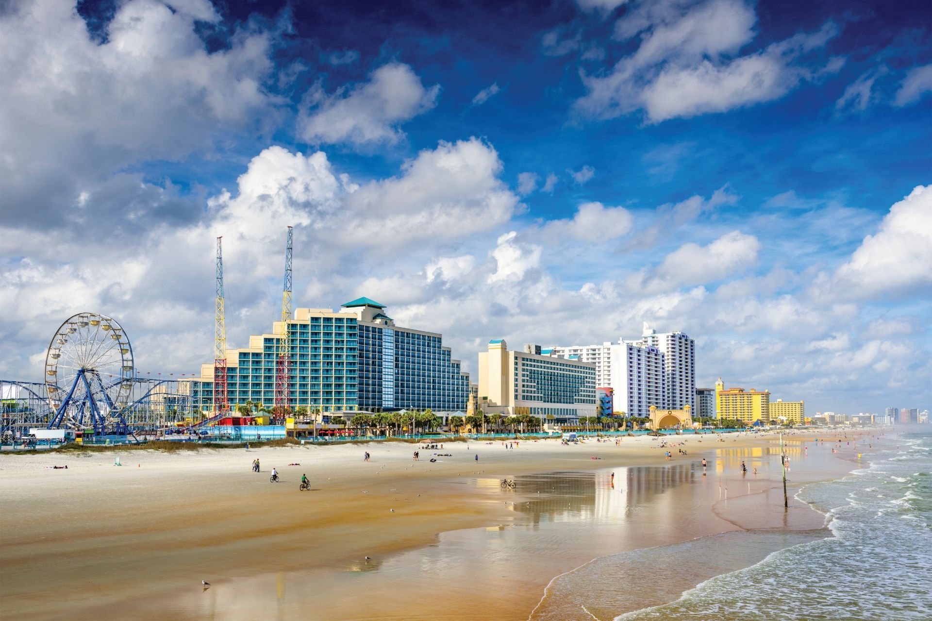 Beachfront city skyline with a Ferris wheel, hotels, and people on a sandy shore under a bright blue sky
