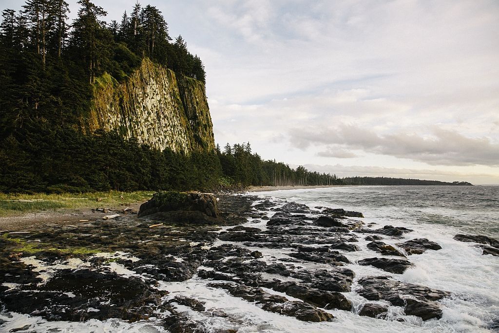 Rocky beach with a forested cliff on the left under a cloudy sky.