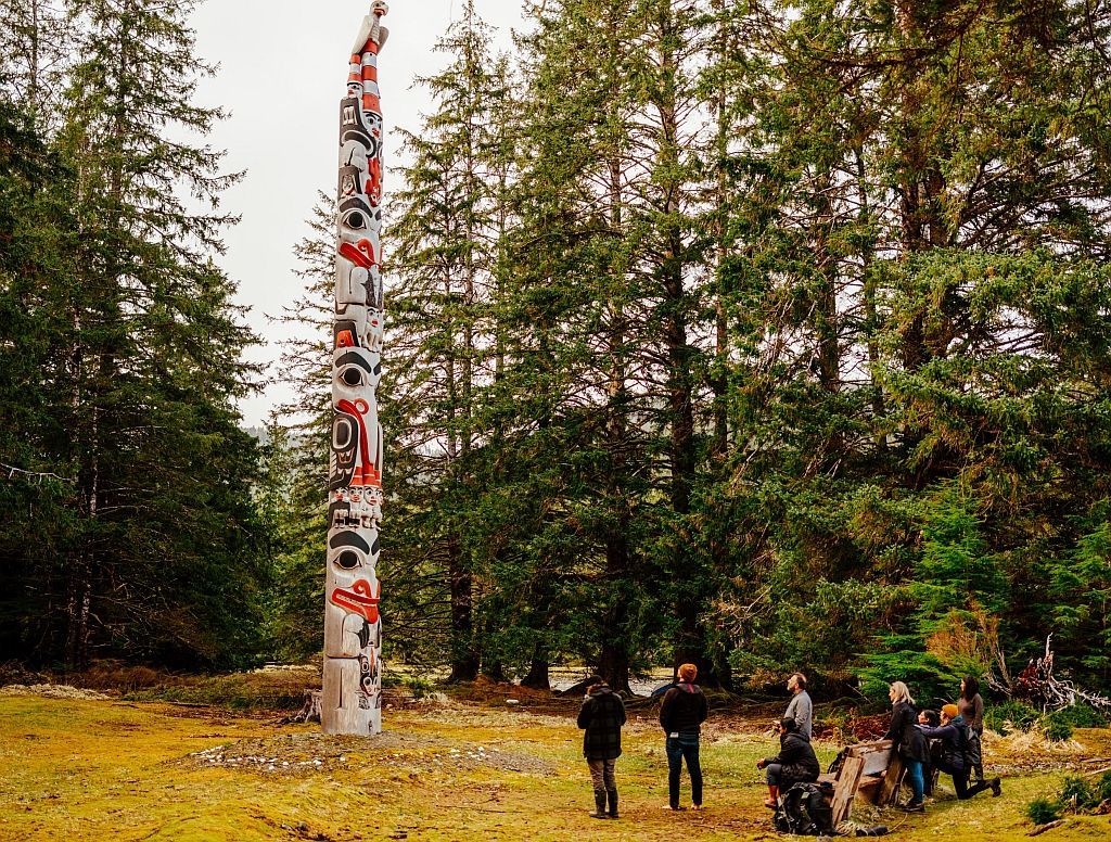 Totem pole in a forest, people observing. Tall pole with carvings, surrounded by trees and overcast sky.