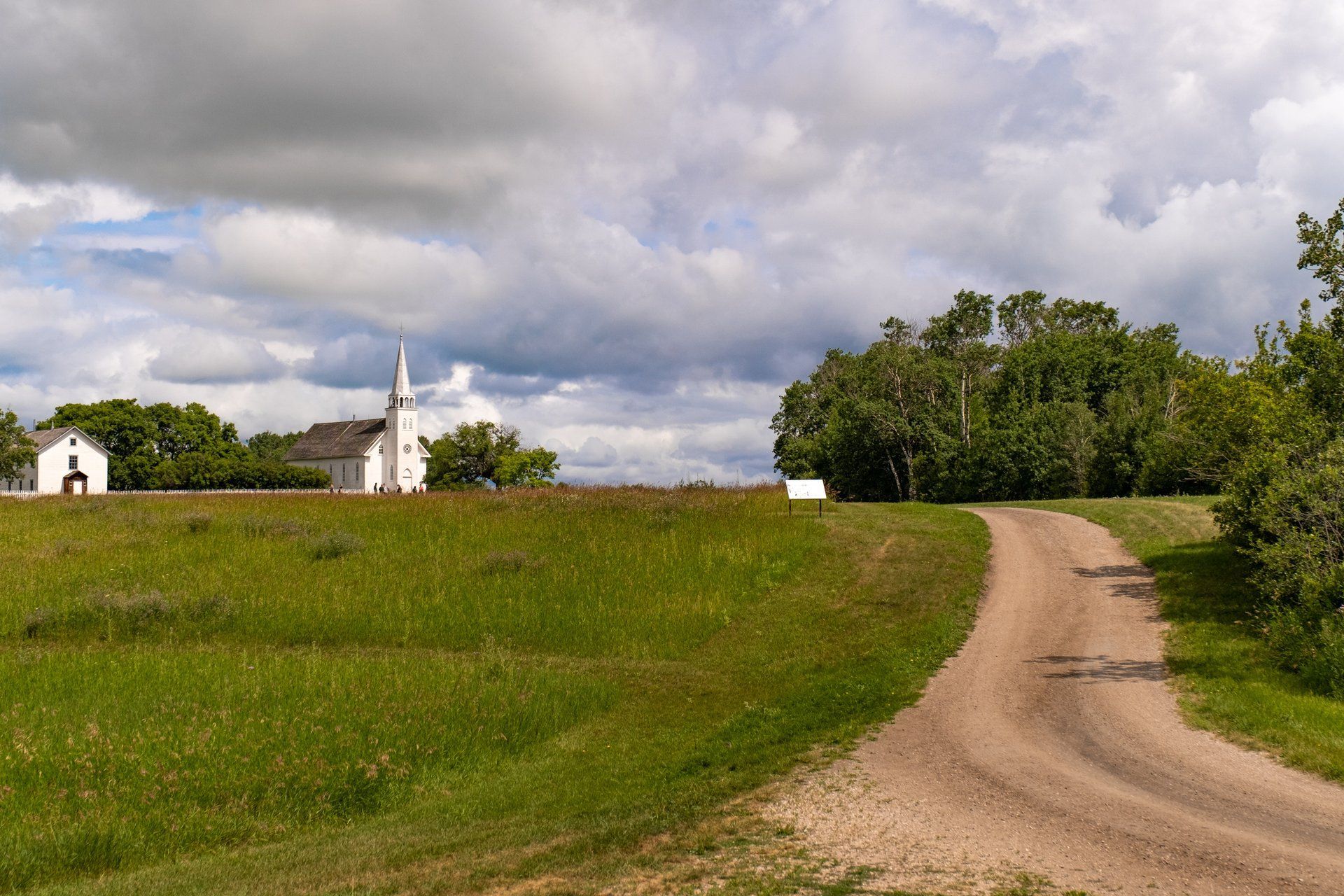 Dirt road leading to a white church with a steeple, on a green hill, under a cloudy sky.