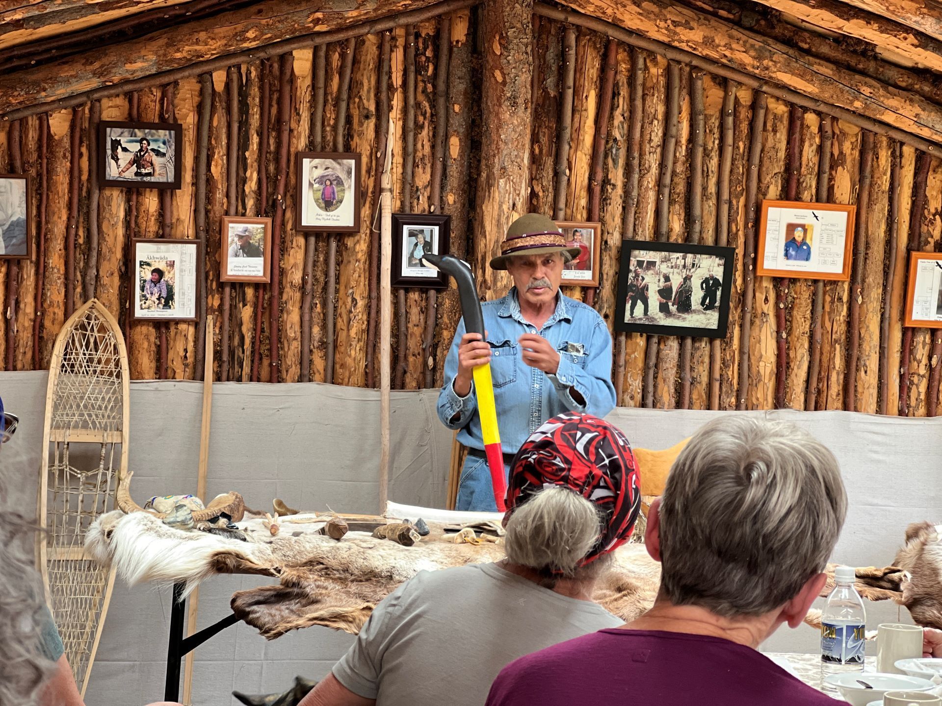 Man in a hat demonstrating a tool to an audience in a wooden building, with artifacts on a table.