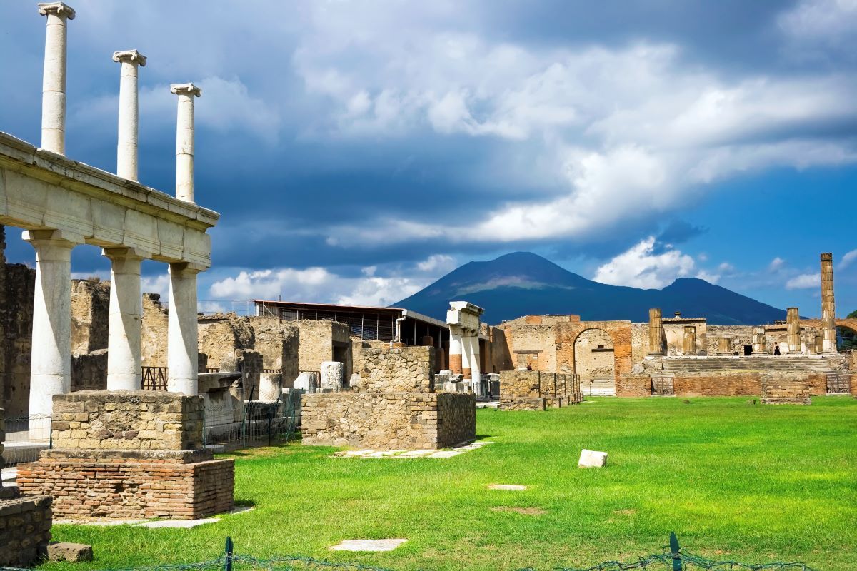 Ruins of Pompeii with columns in the foreground, green grass, and Mount Vesuvius in the background under a cloudy sky.