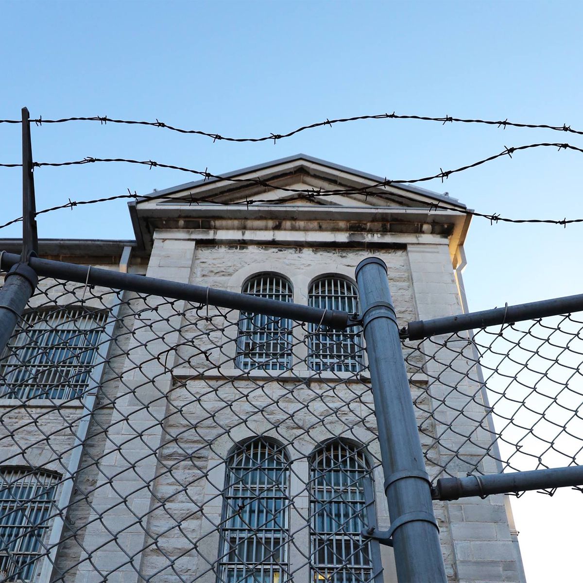 Prison building behind a chain-link fence topped with barbed wire against a blue sky.