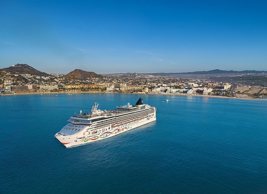Scenic view of a blue bay surrounded by green hills, with boats and pink flowers in the foreground under a blue sky.
