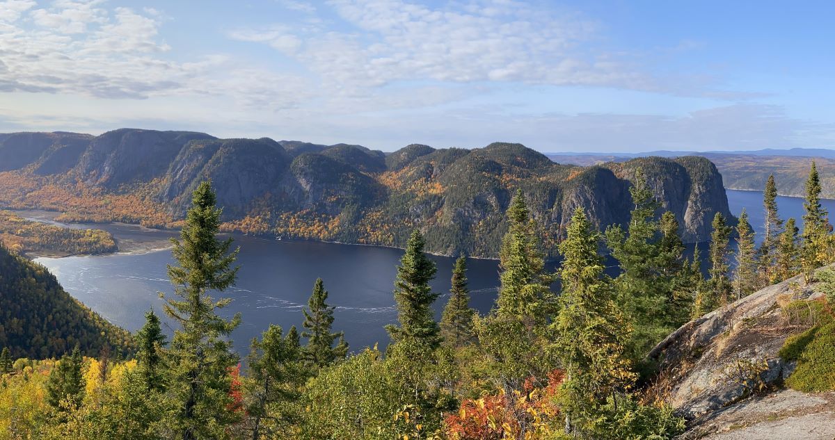 Scenic view of a lake surrounded by forested mountains, some with autumn foliage, under a blue sky.