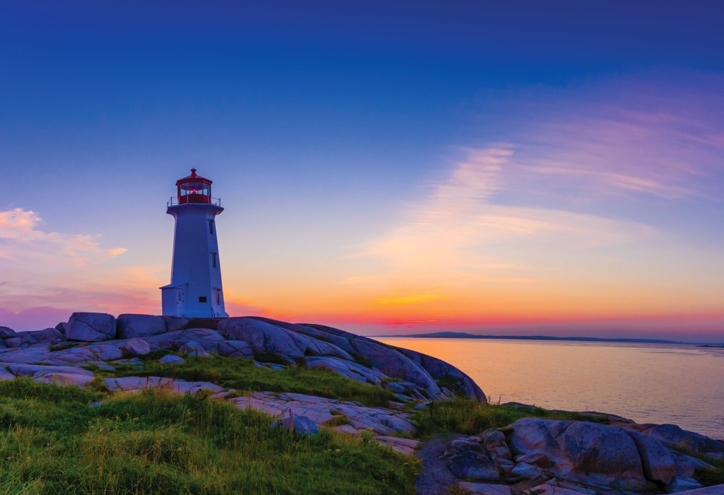 Lighthouse on rocky shore at sunset; orange and purple sky, calm ocean.