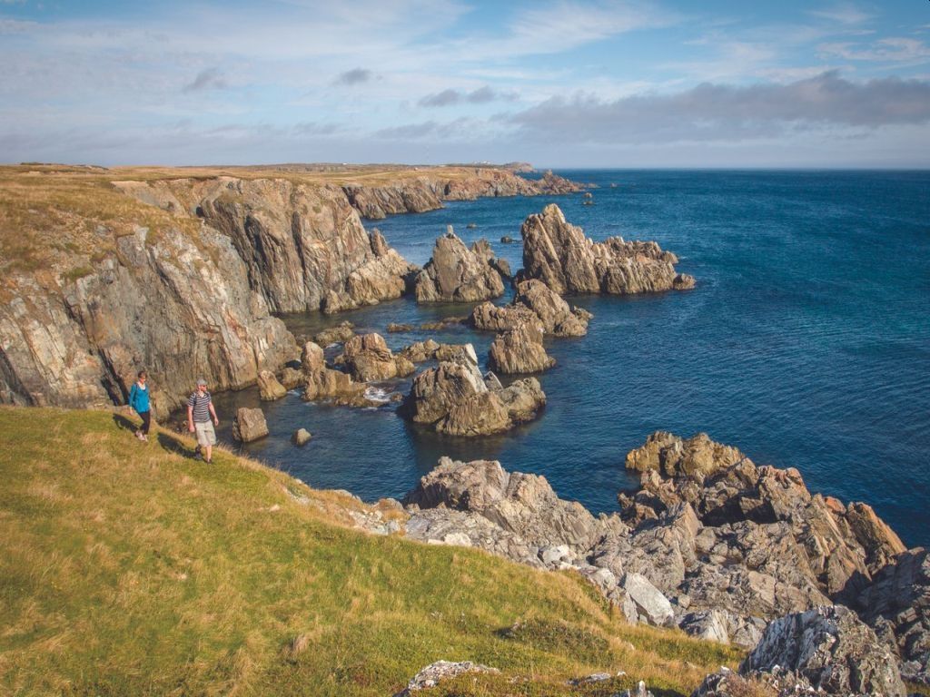 Rocky sea cliffs and blue ocean, with a few hikers on a grassy overlook.