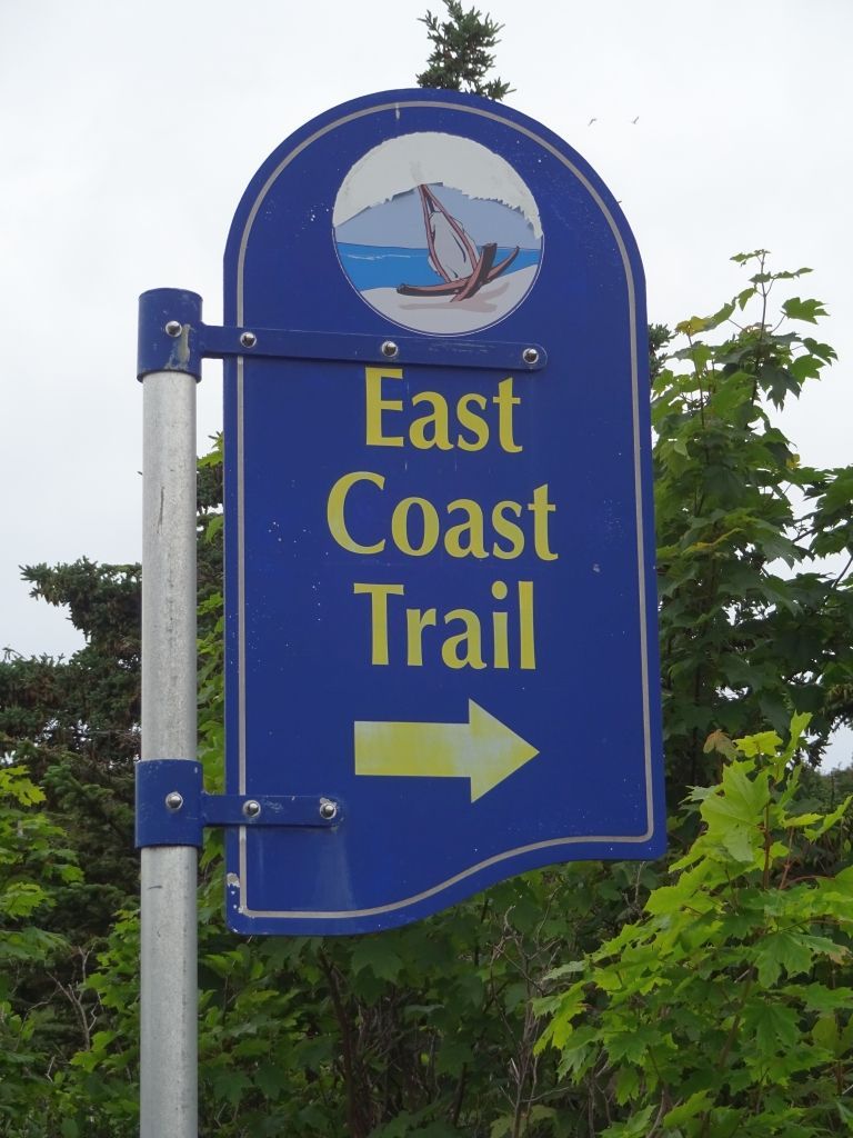 A blue trail sign for the East Coast Trail featuring a yellow right-pointing arrow, mounted on a pole against green trees.