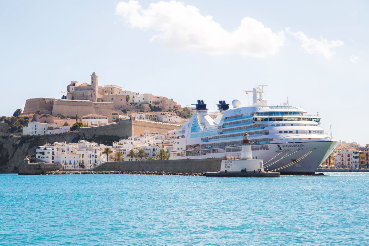 Cruise ship docked in Ibiza harbor, white buildings and castle on a hill under a sunny sky.