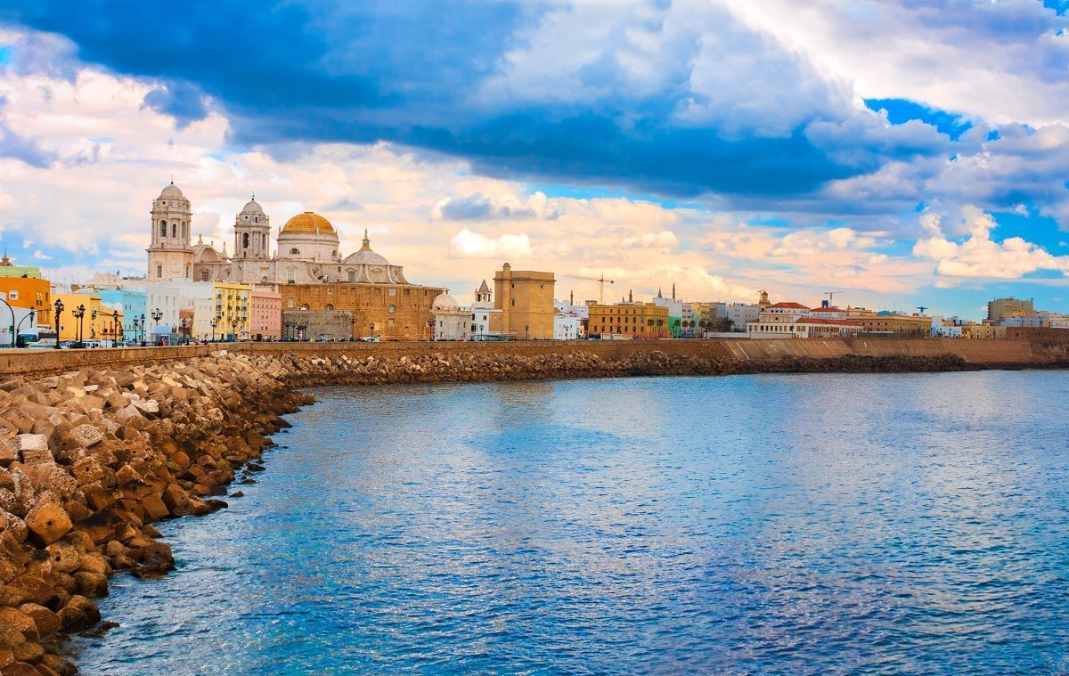 Coastal cityscape with church, buildings, and water under a cloudy sky.