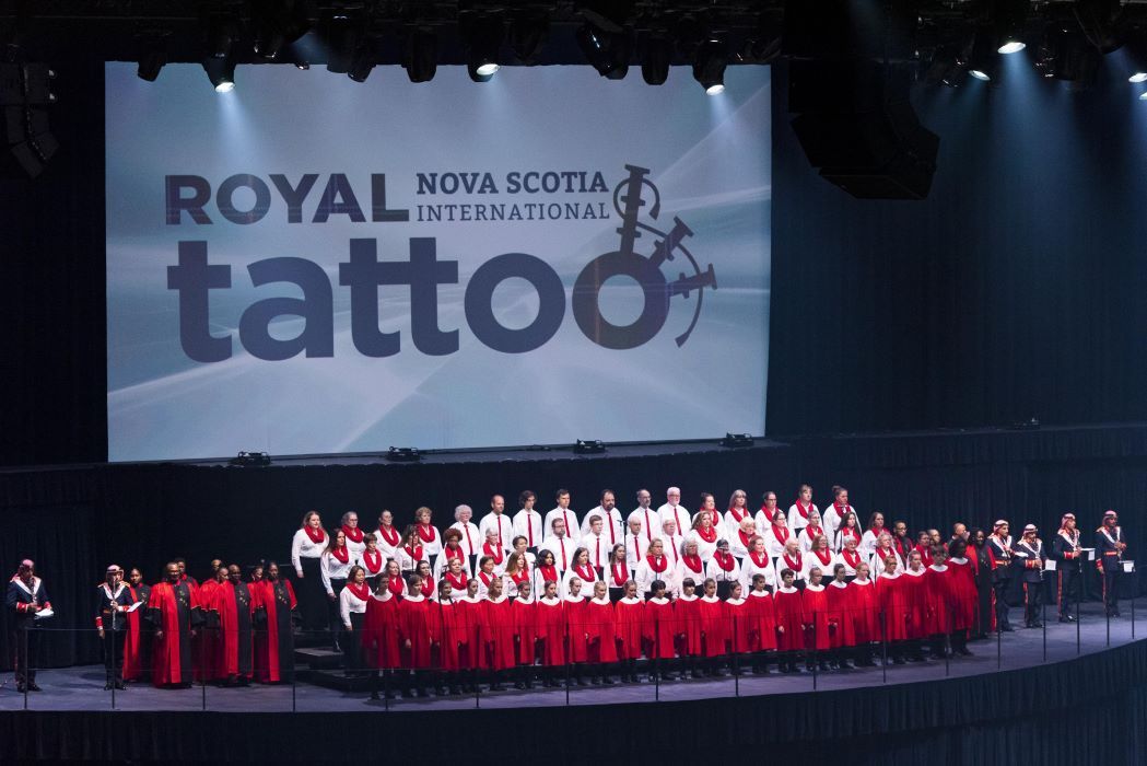 Scottish dancers in kilts perform with arms raised, accompanied by a bagpipe band on a stage.