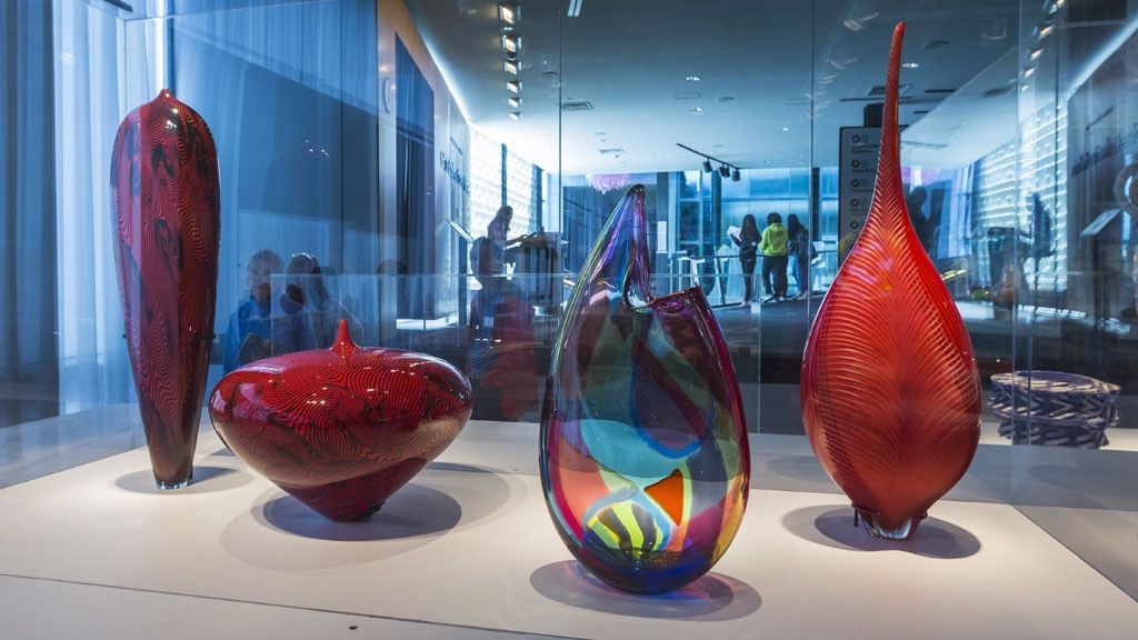 Glass art display in a gallery, showcasing various colorful abstract shapes, with window to street in background.