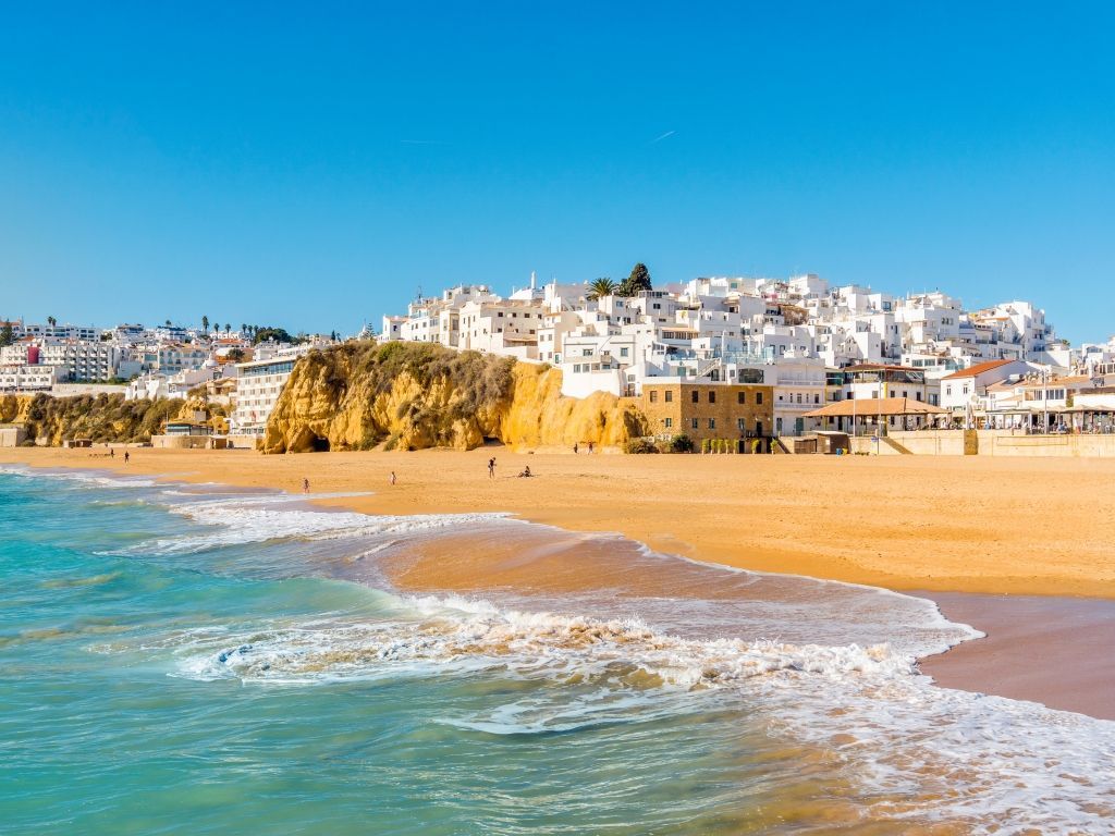Beach scene with golden sand, turquoise water, and white buildings on a cliff under a clear blue sky.