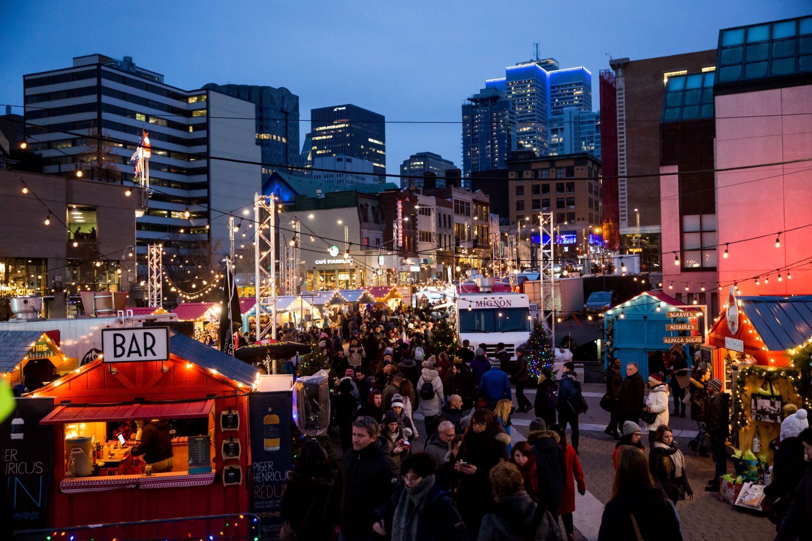 A bustling outdoor market at dusk. People gather around food stalls with city buildings in the background.