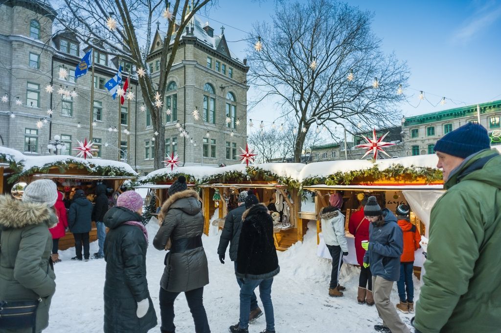 People at a winter market with illuminated stalls, decorated with lights, with buildings in the background.