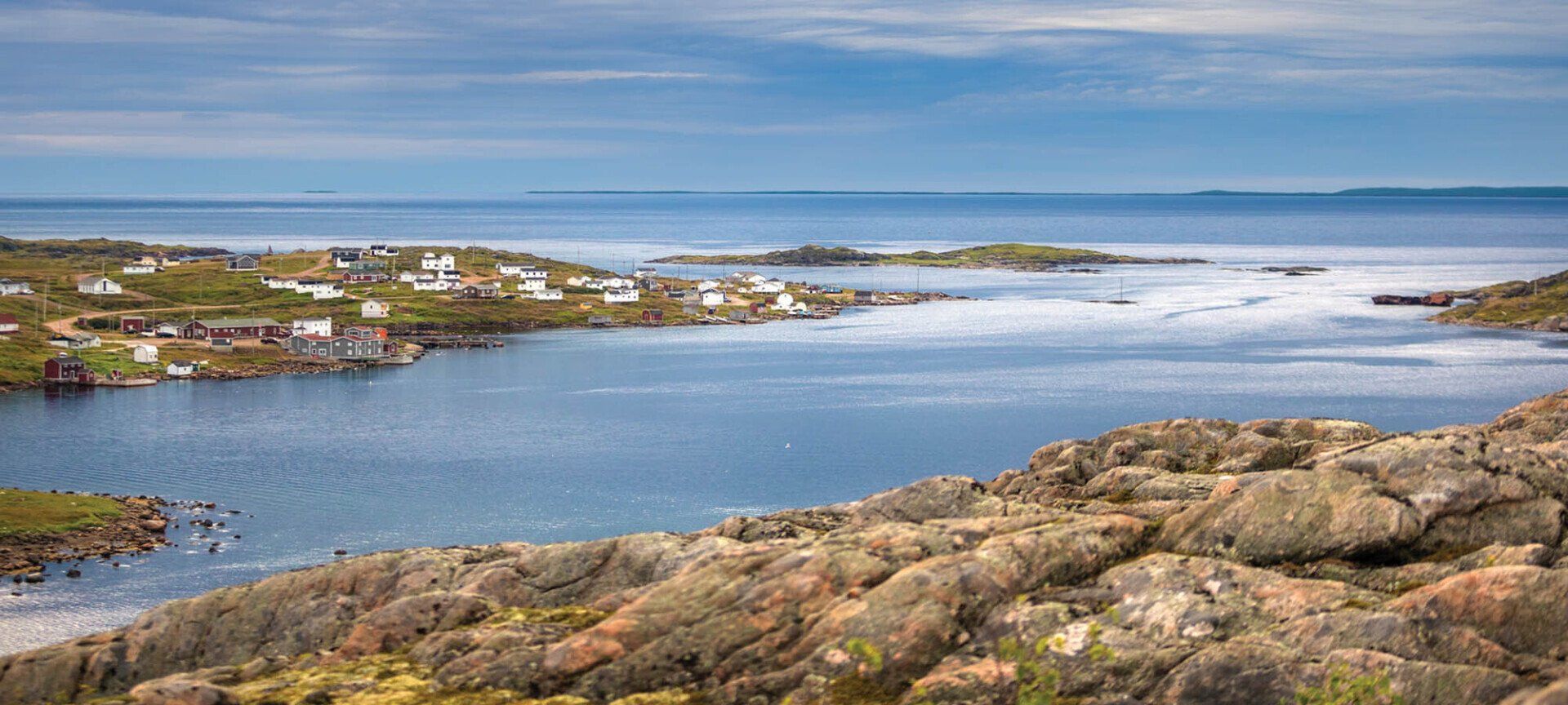 Coastal village nestled by a calm blue sea. Rocky foreground, cloudy sky.