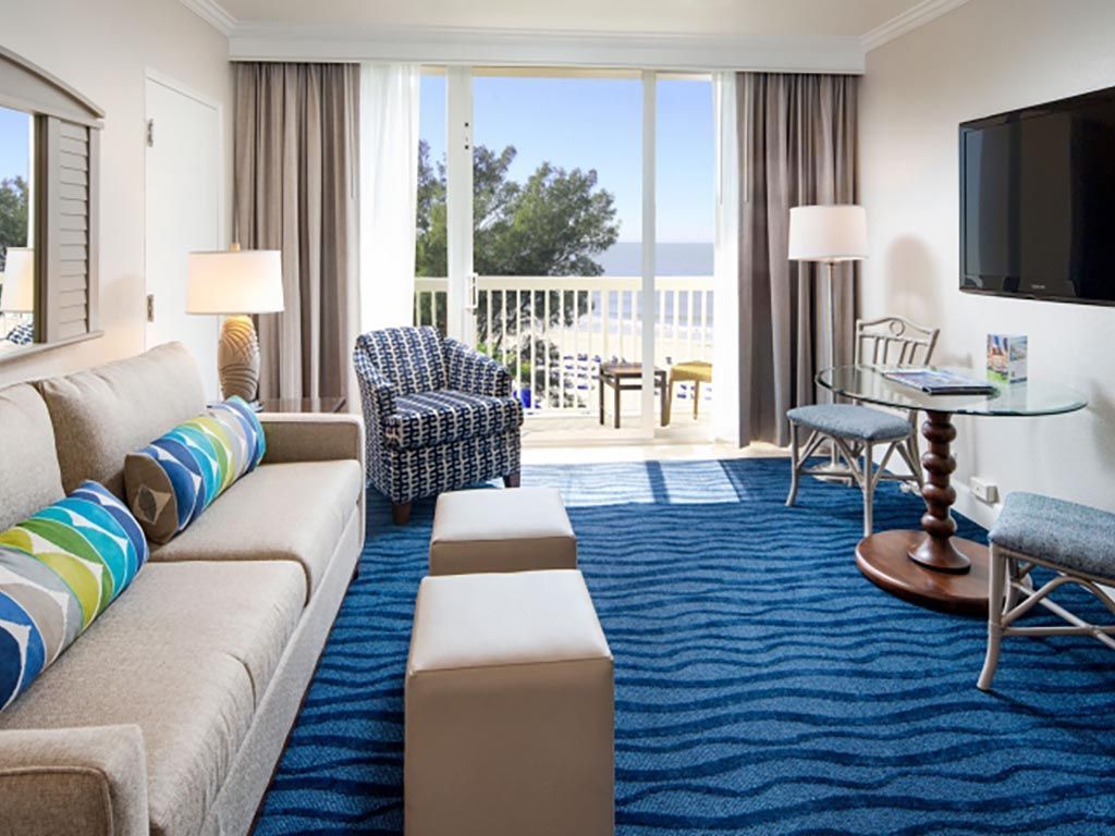 Bright hotel room with ocean view, sofa, armchair, table, and blue patterned carpet.